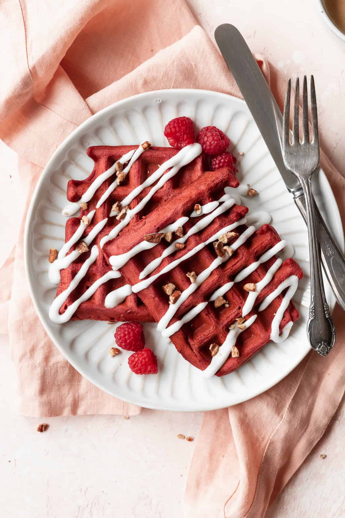 A closeup shot of a white plate of red velvet waffles with cream cheese glaze and raspberries.