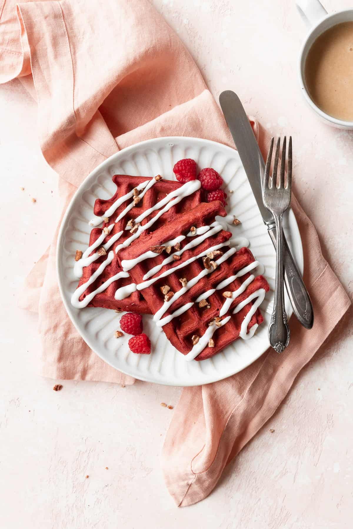 An overhead shot of a white plate of red velvet waffles with cream cheese drizzle and pecans.