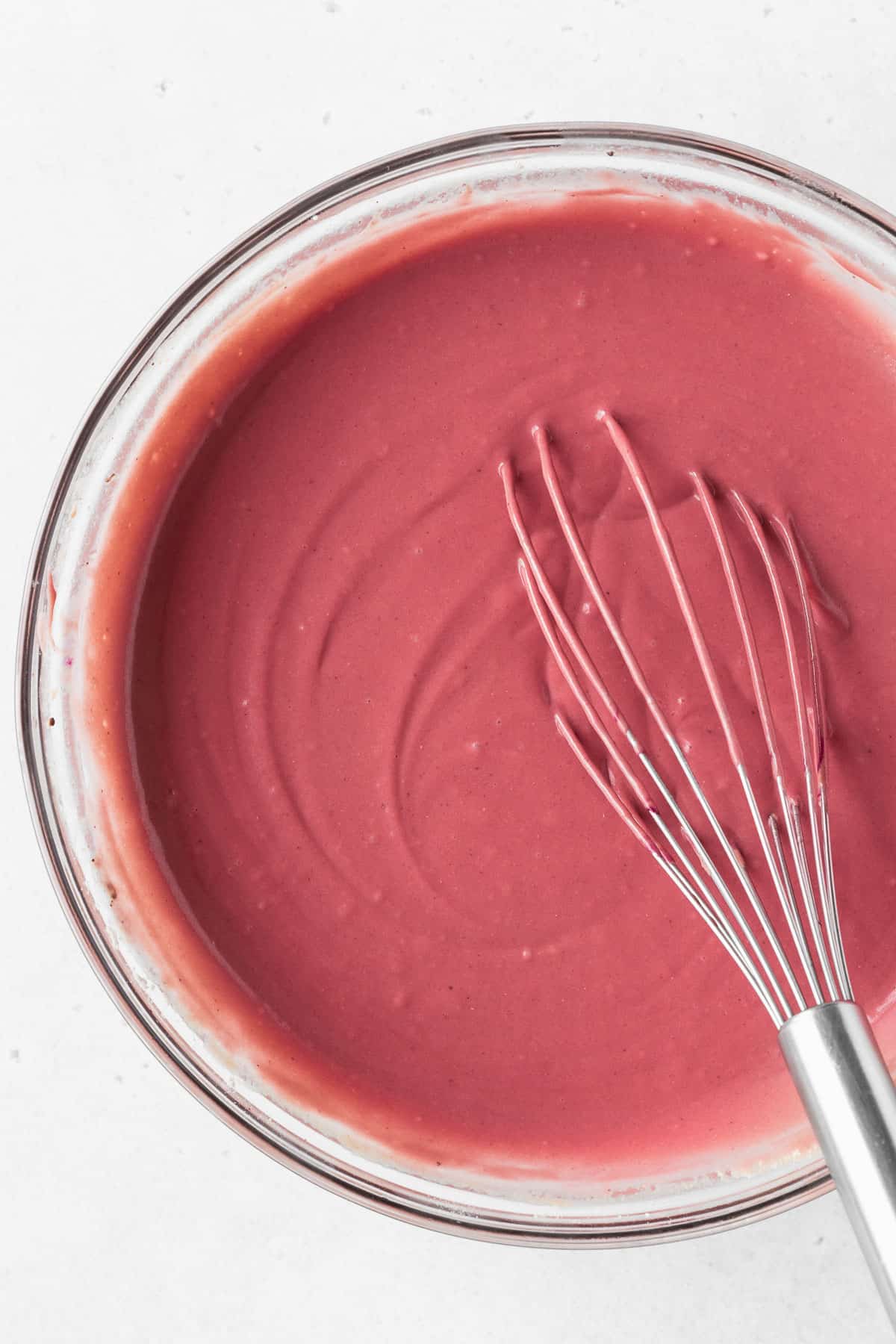 A closeup overhead shot of a glass mixing bowl of red velvet waffle batter with a whisk.