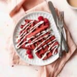 A square shot of a white plate of red velvet waffles with cream cheese drizzle and pecans.