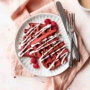 A square shot of a white plate of red velvet waffles with cream cheese drizzle and pecans.