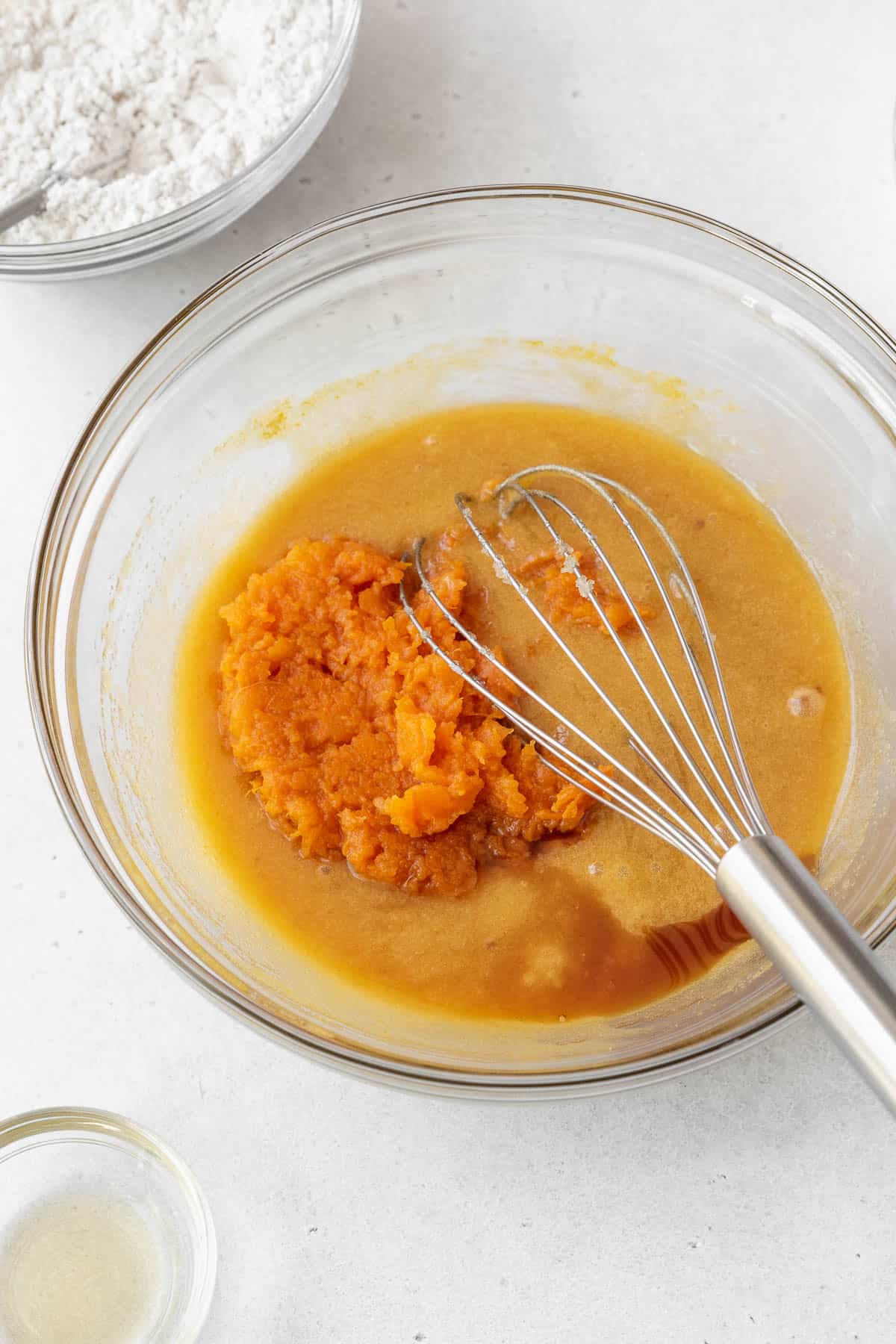 Adding mashed sweet potato and vanilla extract to a glass bowl on a white surface.