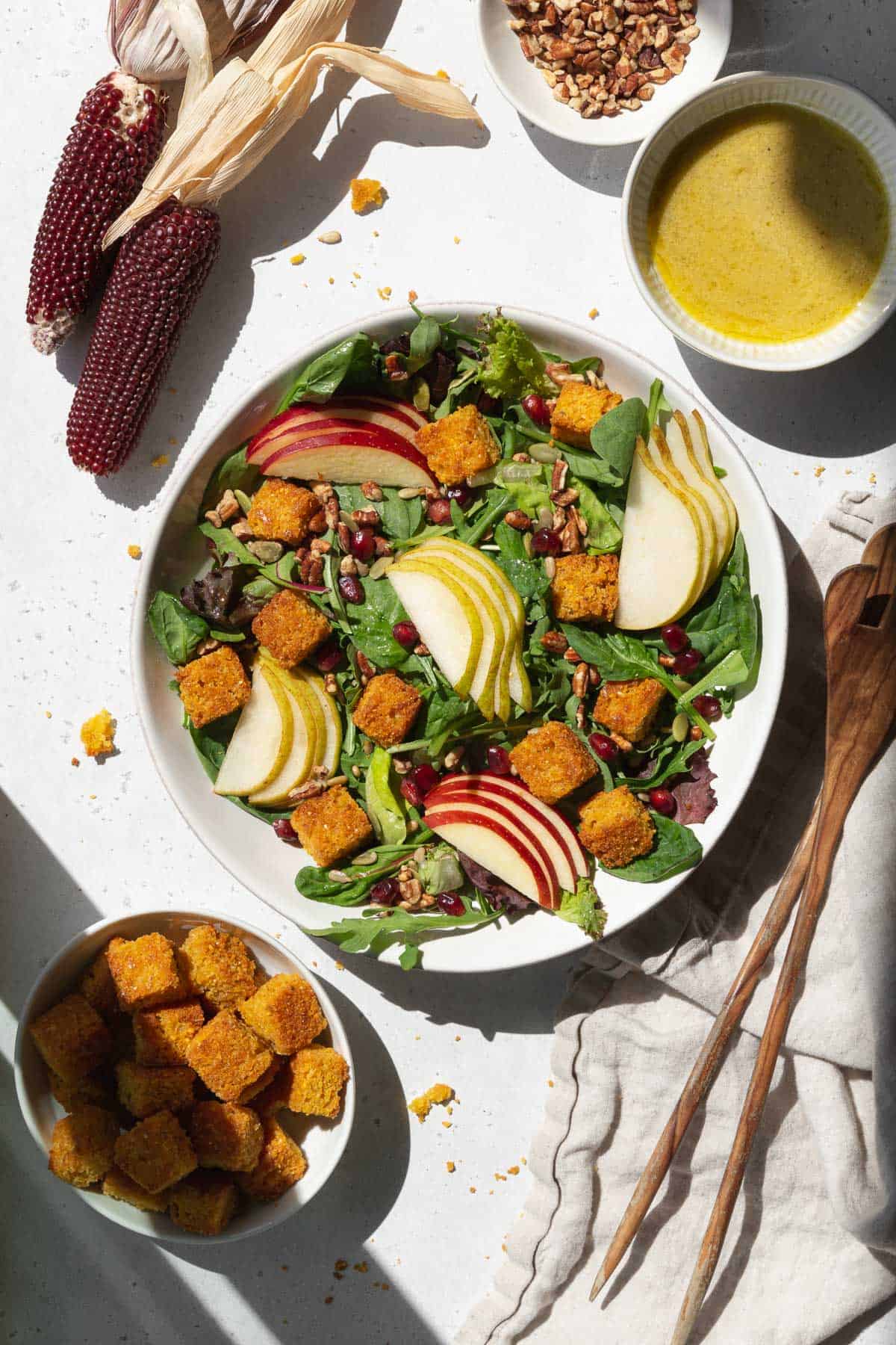 A spread of salad with cornbread croutons, pears, apples, dressing, and salad tongs.