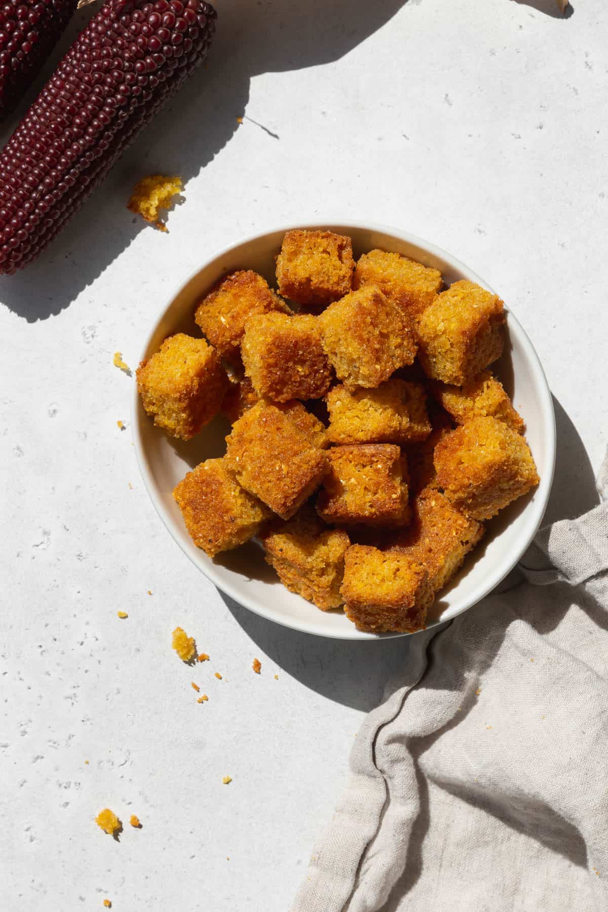 An overhead shot of a small bowl of cornbread croutons with a napkin and a stalk of heirloom corn.