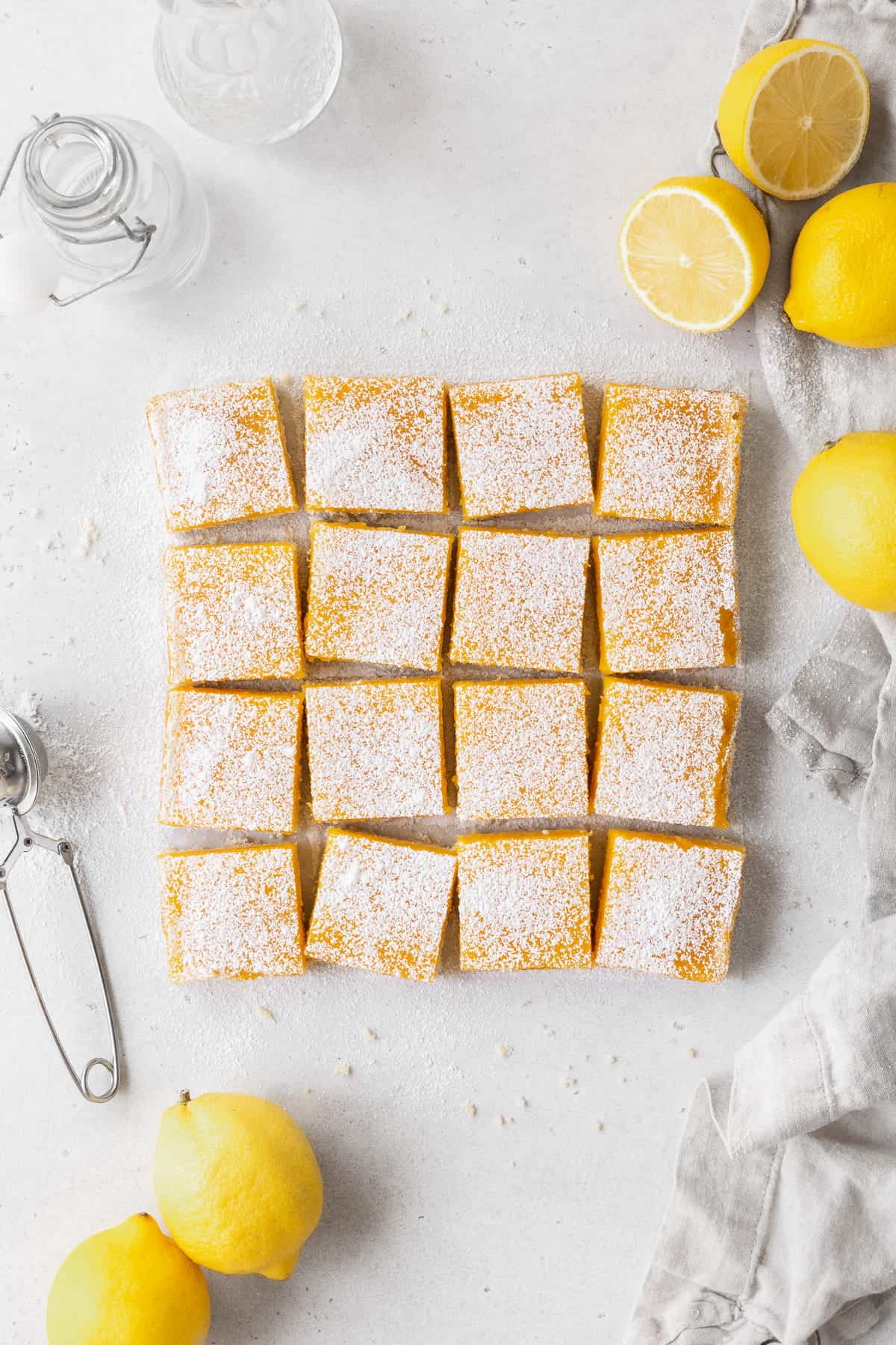 A pan of dairy free lemon bars cut up on the counter with halved lemons, powdered sugar, and a napkin.