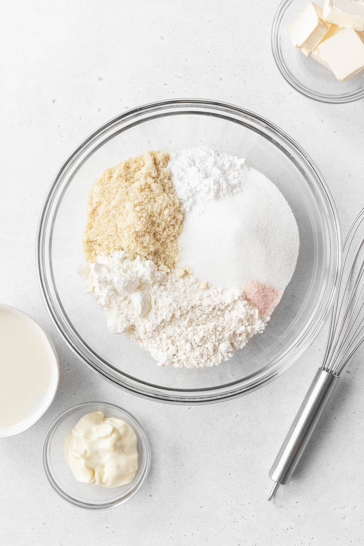 All of the dry ingredients for gluten-free and dairy-free tres leches cake in a glass mixing bowl with a whisk. The other cake ingredients sit beside the mixing bowl.