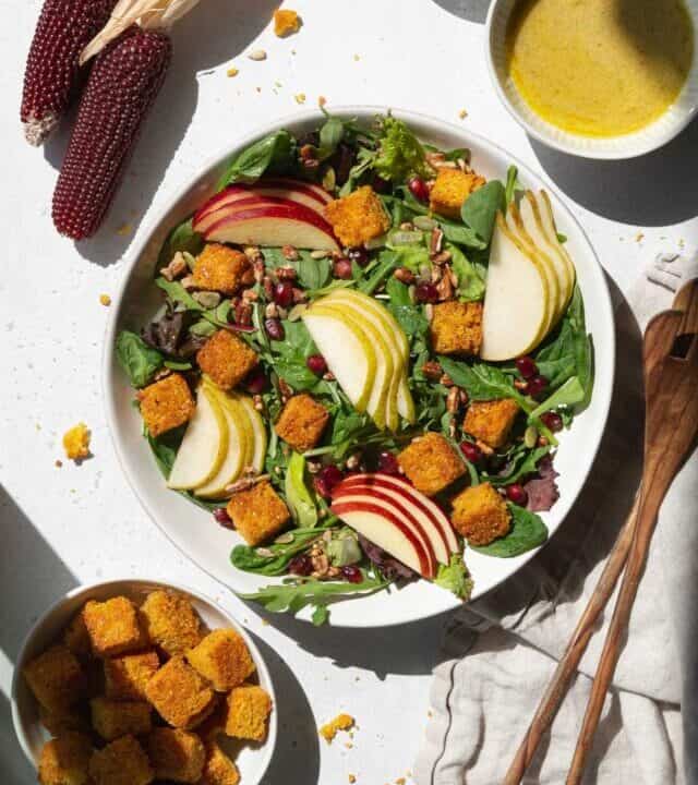 A spread of salad with cornbread croutons, pears, apples, dressing, and salad tongs.