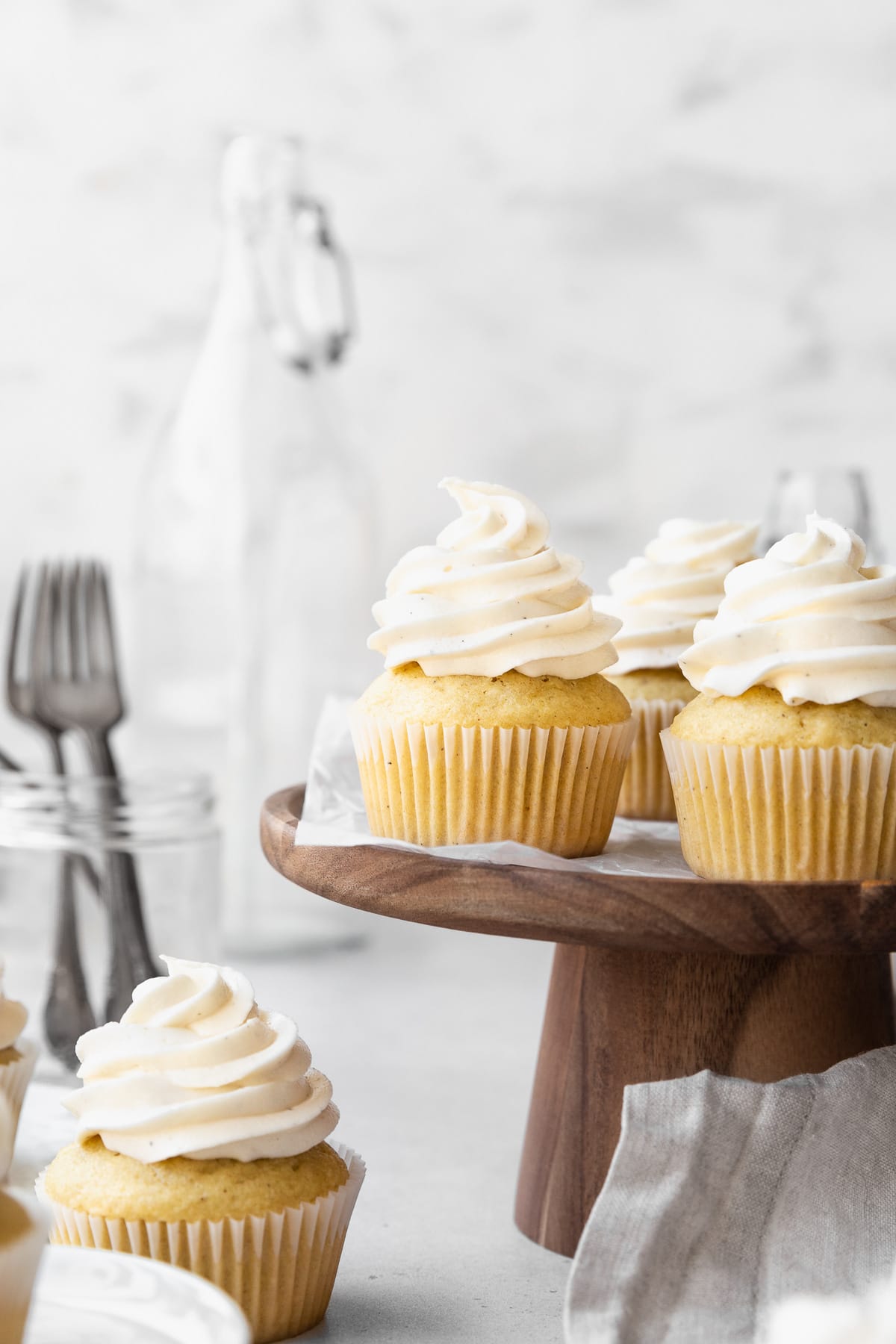Side on shot of dairy-free vanilla cupcakes on a wooden cake stand with a jar of forks and a large jug in the background.
