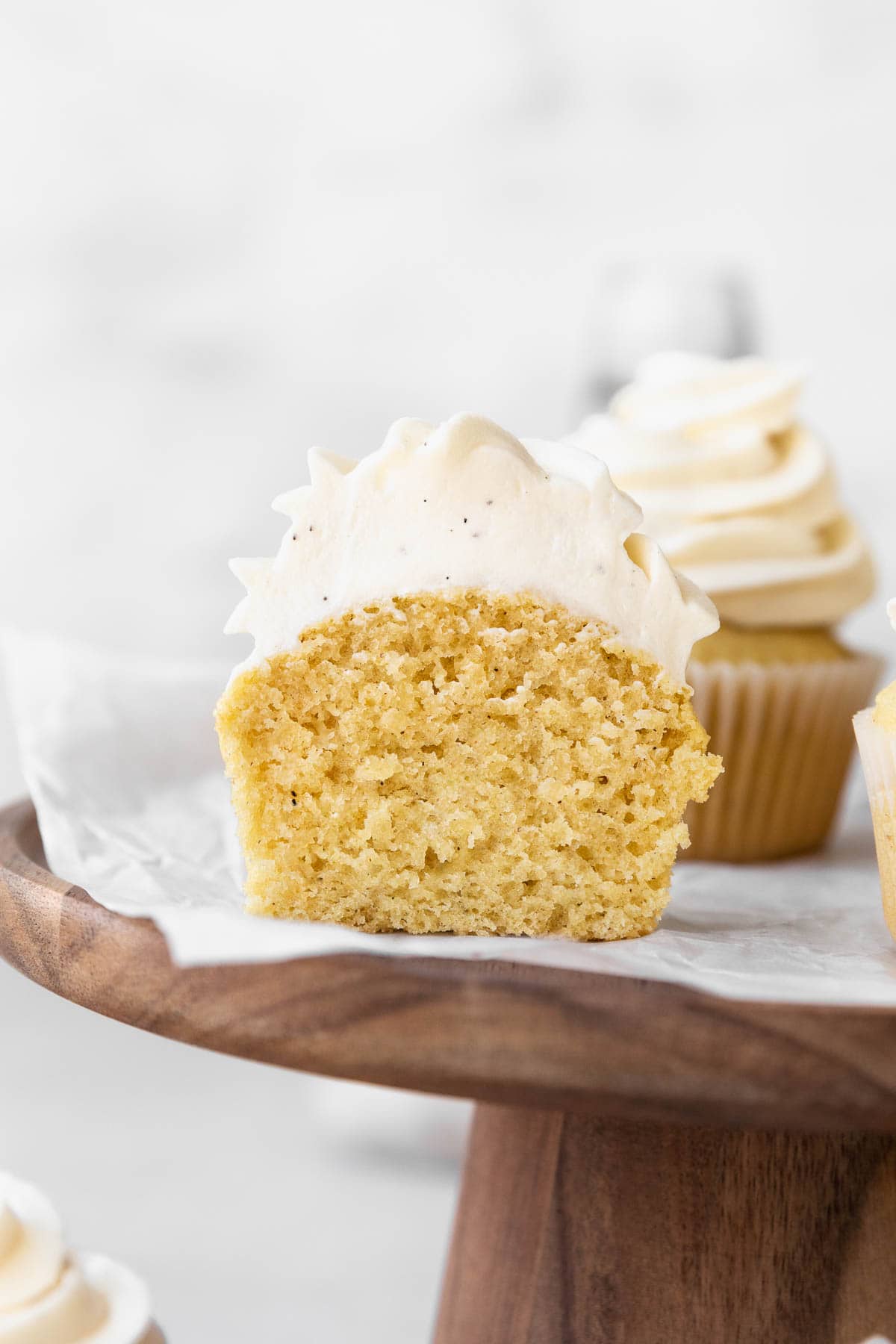 A dairy free vanilla cupcake topped with dairy free vanilla buttercream on a wooden stand, sliced in half to show the fluffy and tender crumb.