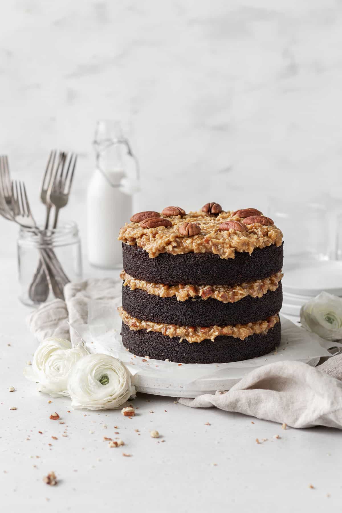 A three-layer gluten-free German chocolate cake on a round wooden board with white flowers beside it, with a milk jug and a jar with forks inside in the background.