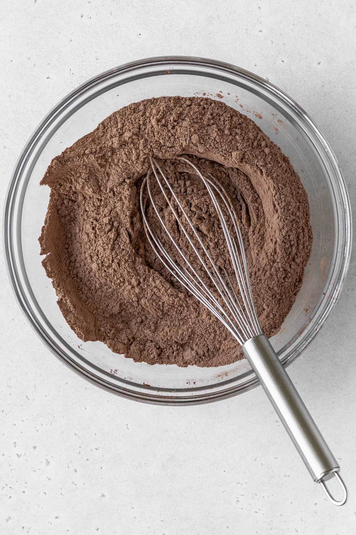 Dry ingredients for a gluten-free chocolate cake in a glass bowl with a silver whisk.