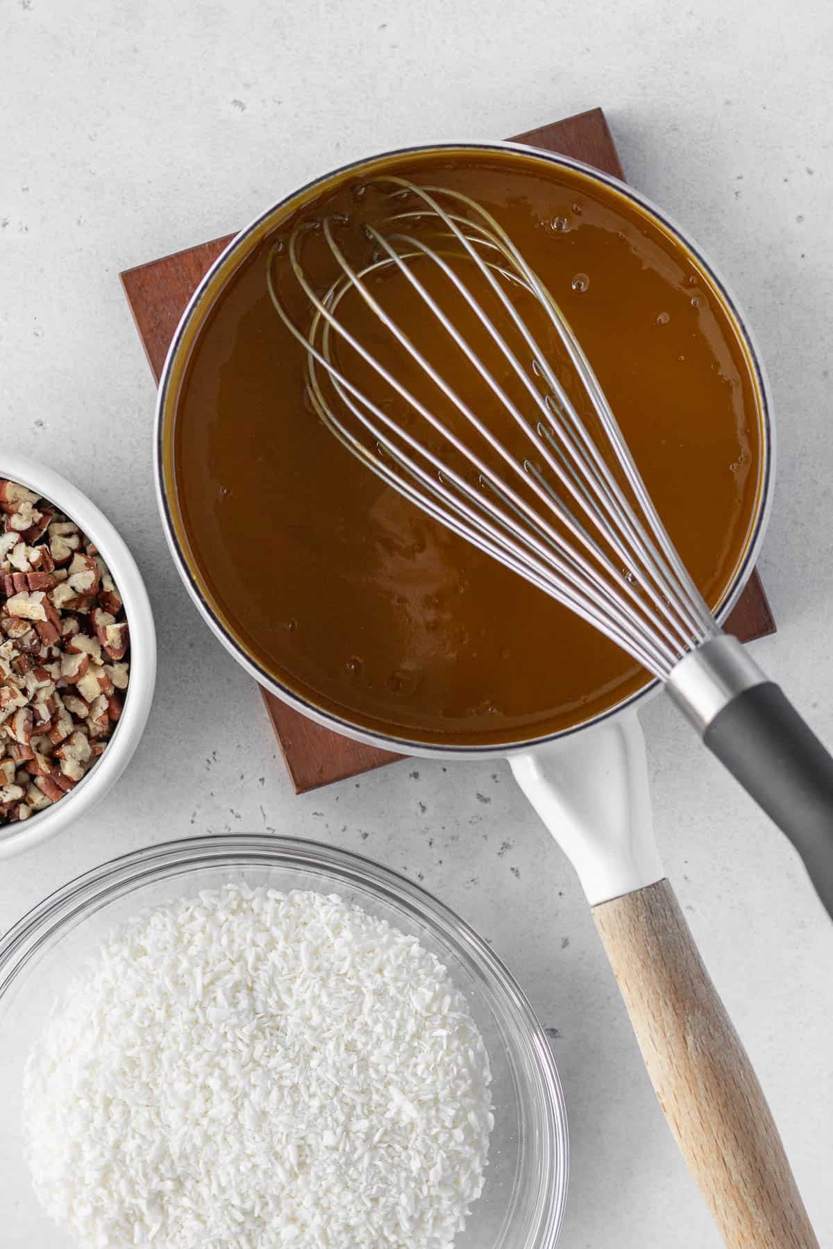 German chocolate frosting mixture being cooked in a saucepan with a bowl of shredded coconut and a bowl of chopped pecans next to it.