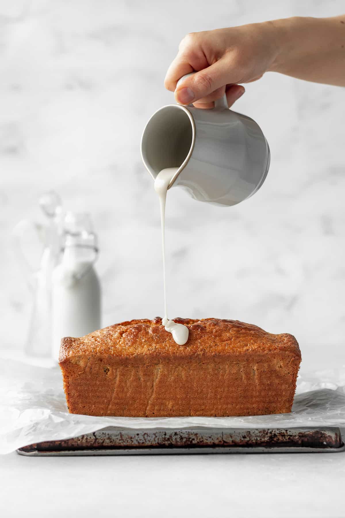 A hand holding a ceramic jug of lemon icing being poured onto a loaf of gluten free lemon poppy seed cake.