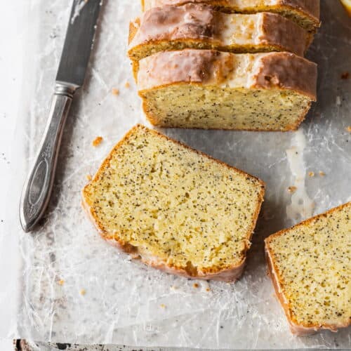A slice of gluten free lemon poppy seed cake laying on parchment paper with a knife next to it and the sliced loaf behind it.