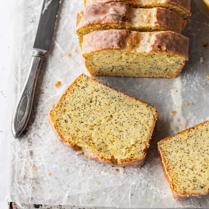 A slice of gluten free lemon poppy seed cake laying on parchment paper with a knife next to it and the sliced loaf behind it.