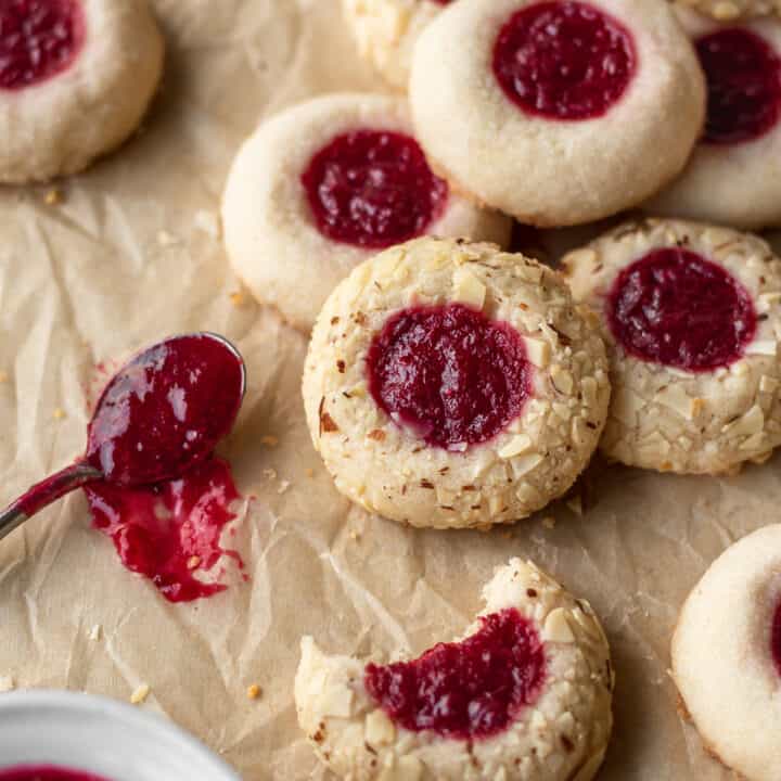A square shot of gluten free thumbprint cookies next to a small spoon of jam on a brown parchment paper.