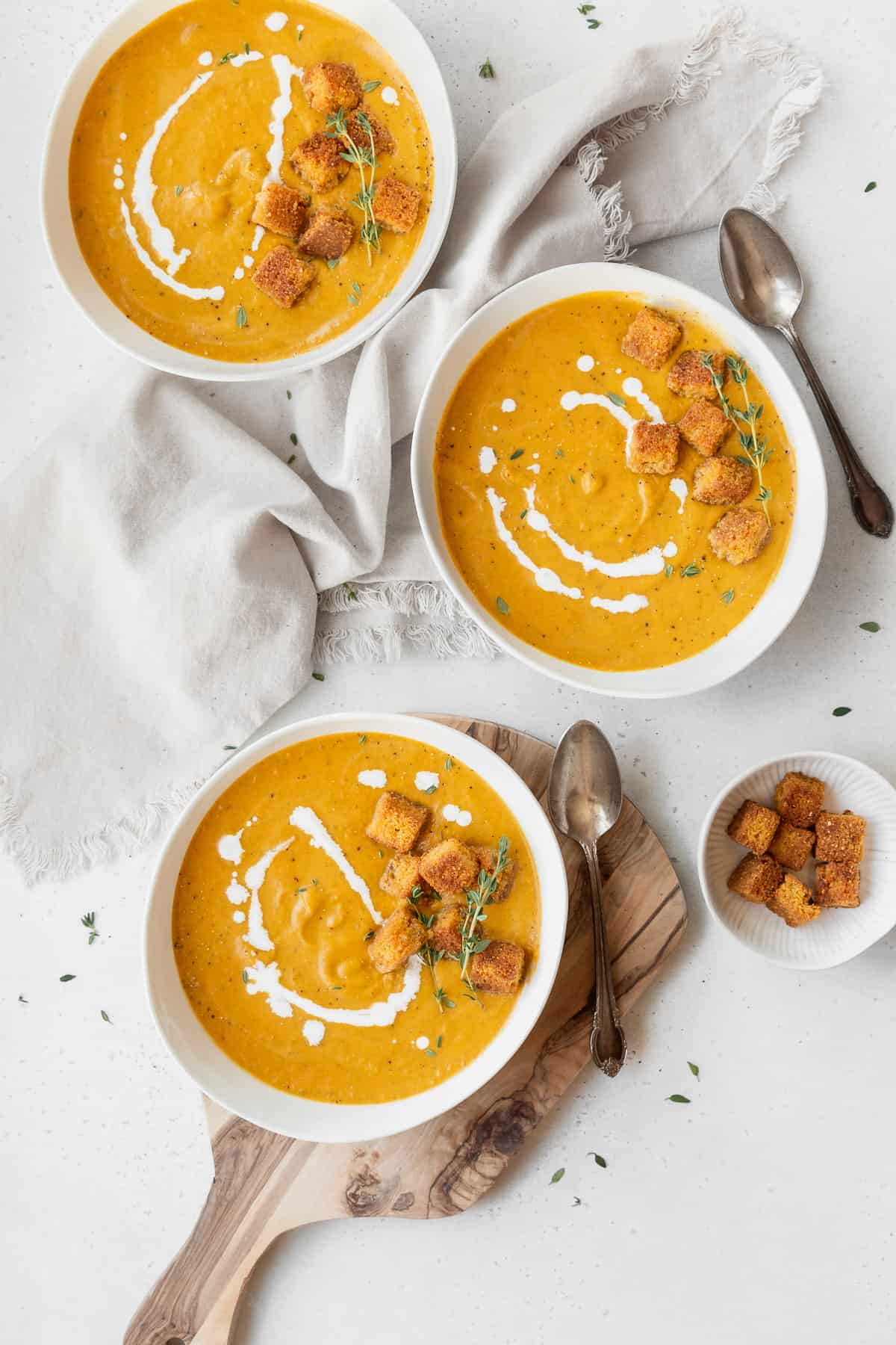 Overhead shot of 3 garnished bowls of vegan pumpkin bisque on a white table with a small bowl of cornbread croutons.