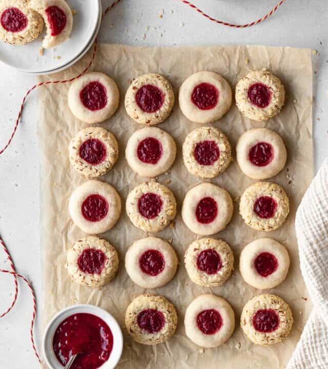 Overhead shot of vegan thumbprint cookies lined up into rows on a piece of brown parchment paper with a small white ramekin full of cranberry sauce.
