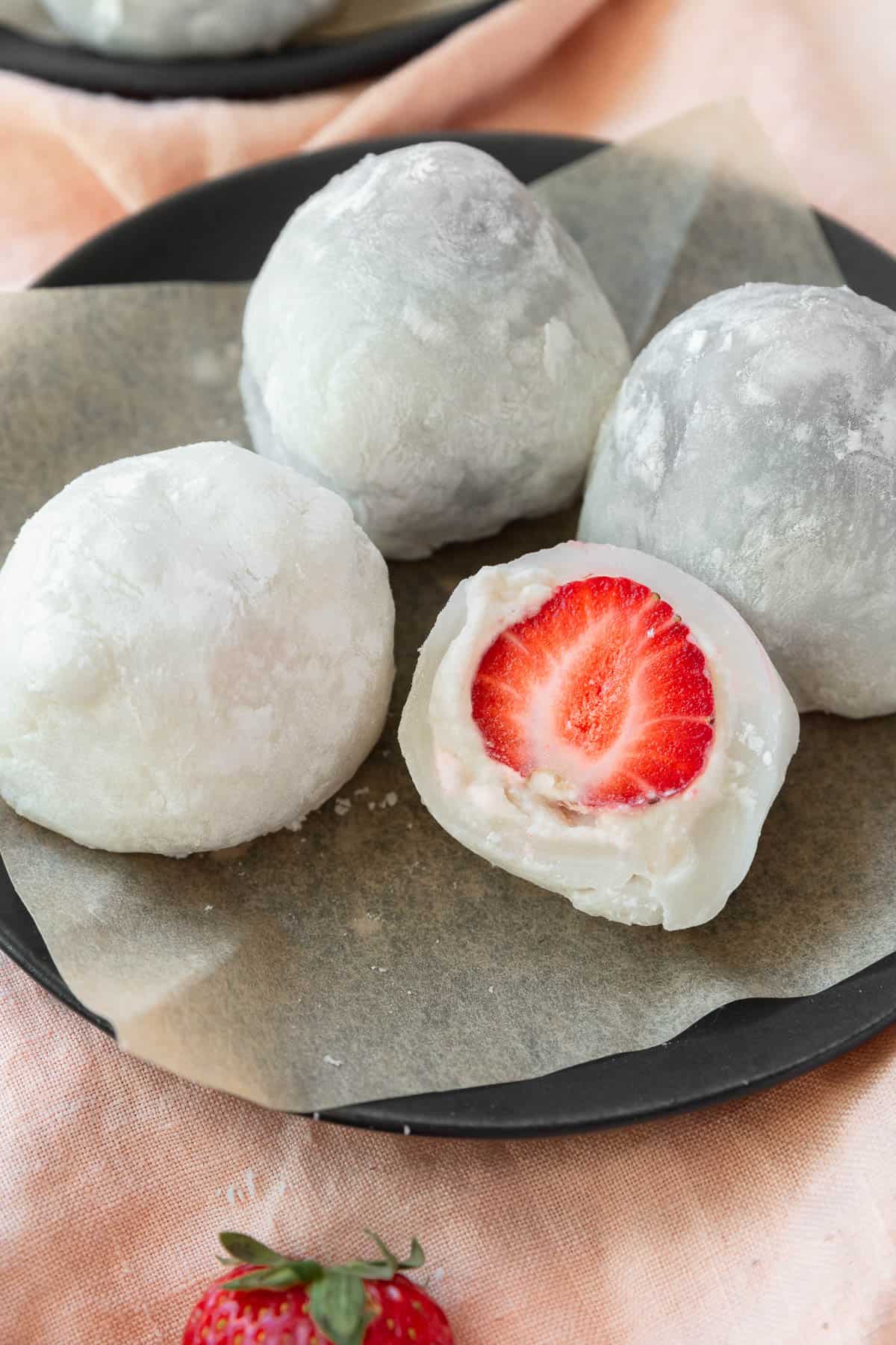 A plate of strawberry mochi with one sliced in half, filled with a strawberry and some cream.