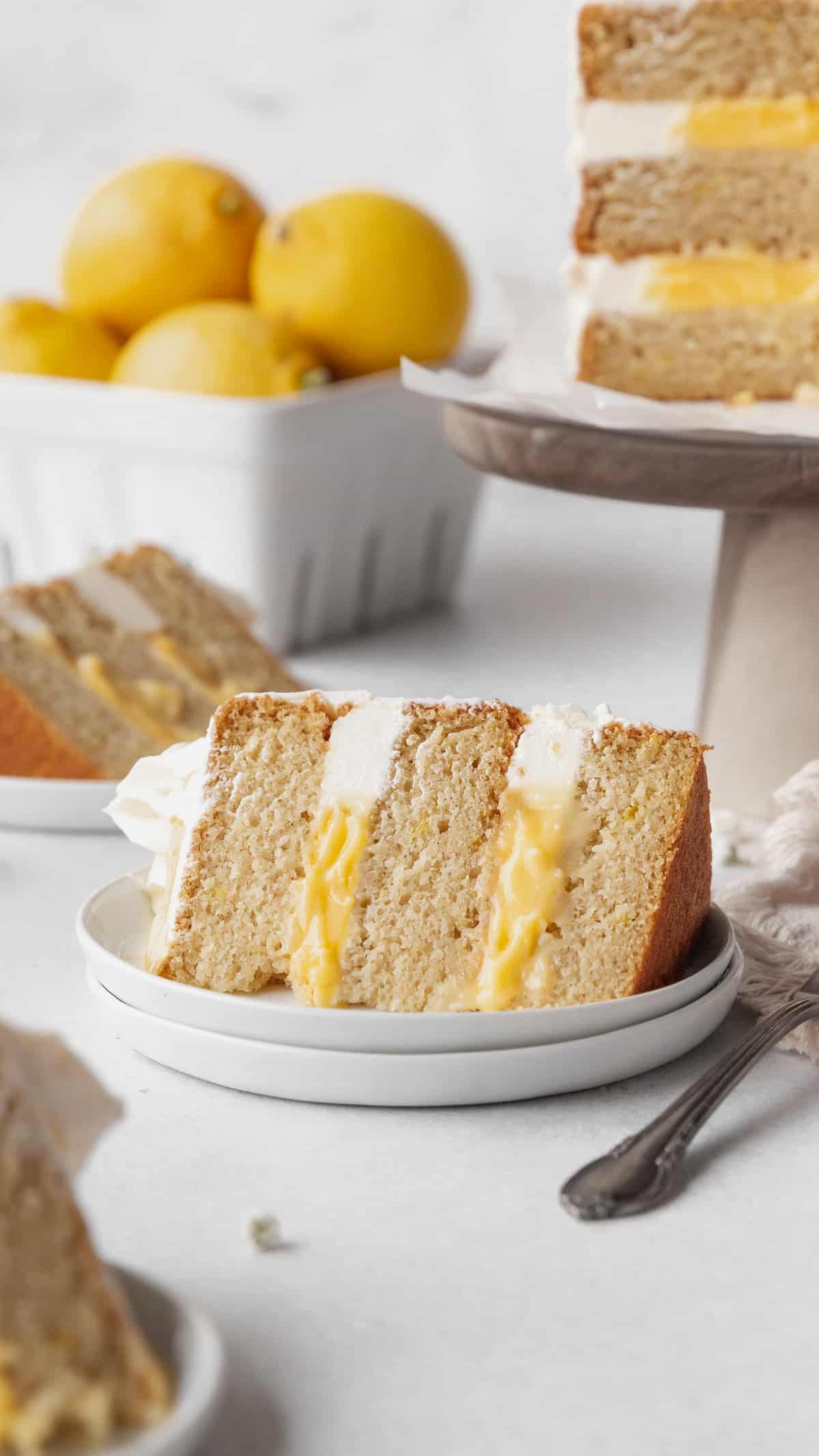 A slice of lemon curd cake on a white dessert plate with a cake stand holding the rest of the cake and a ceramic fruit pint holding a handful of lemons in the background.