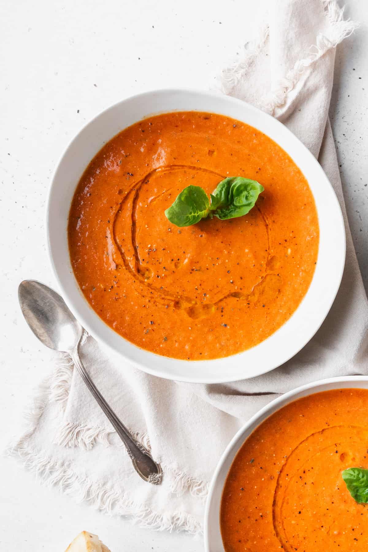 A white bowl of tomato basil soup with a sprig of basil on a linen and a silver spoon placed next to it.