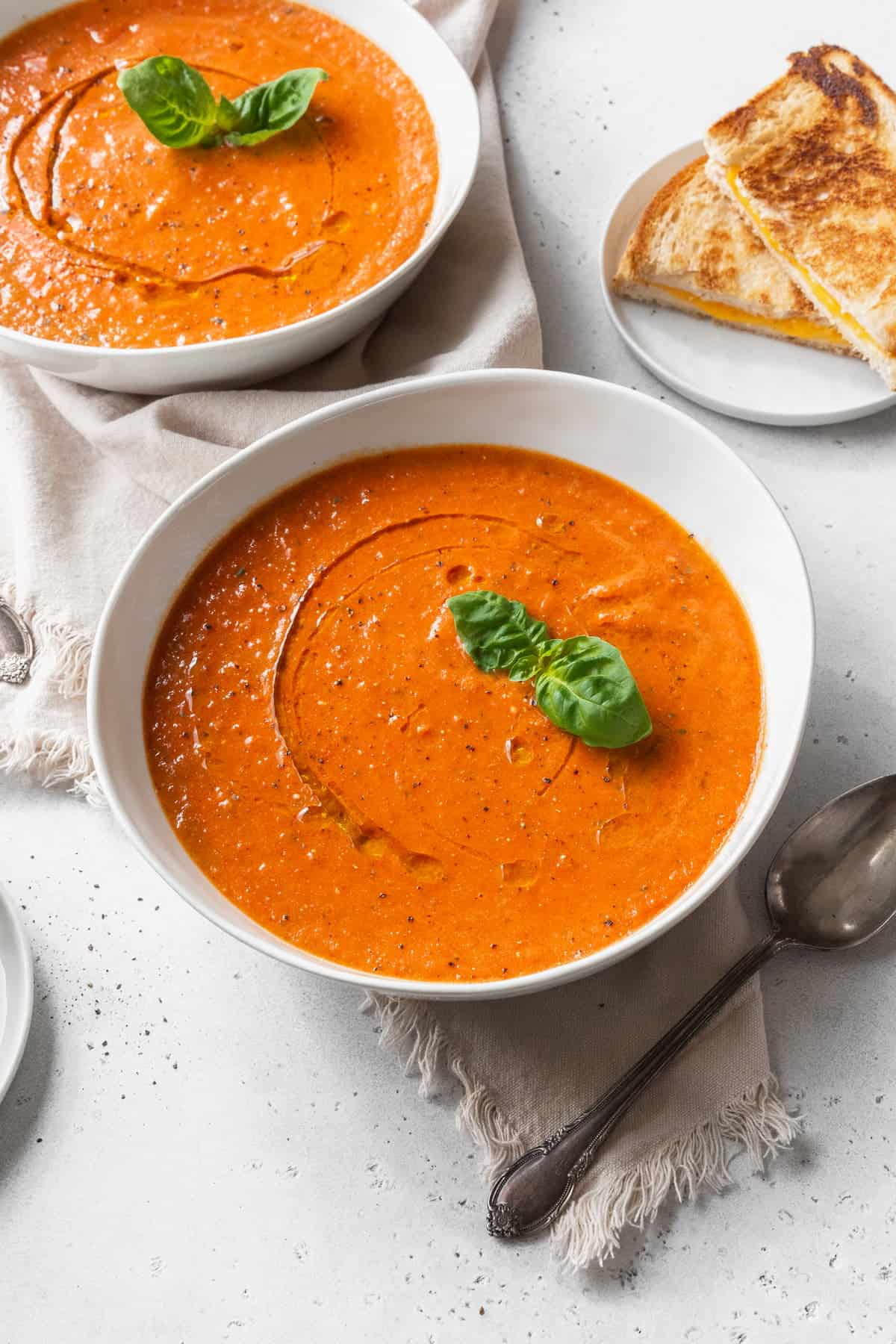 A bowl of vegan tomato soup with a sprig of basil, and a second bowl of soup and some grilled cheese sandwiches in the background.