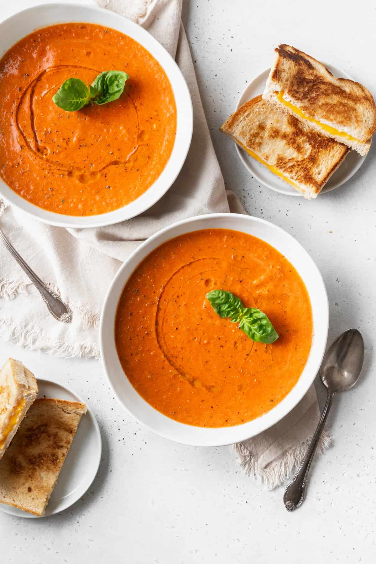 Two bowls of roasted tomato soup next to small plates of halved grilled cheese and spoons on a white surface.