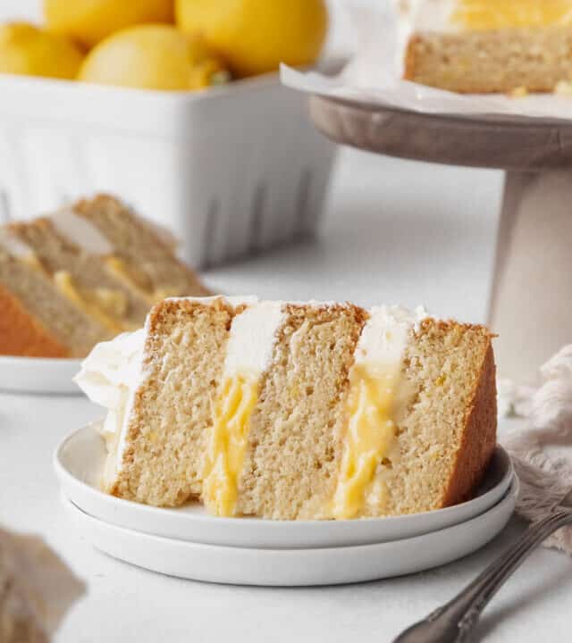 Vertical hero shot of a slice of dairy-free lemon curd cake on a white dessert plate with a cake stand holding the rest of the cake and a ceramic fruit pint holding a handful of lemons in the background.