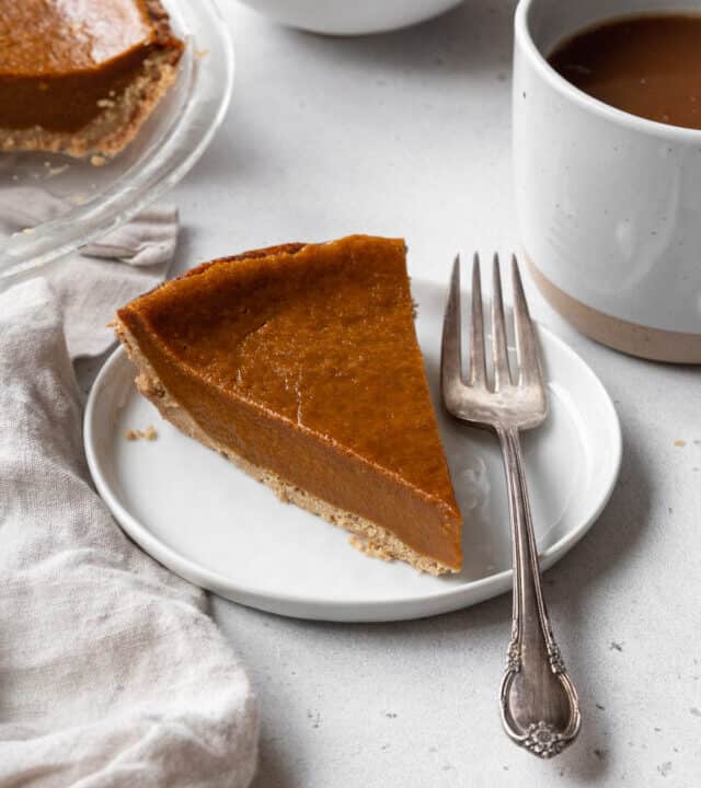 A slice of eggless pumpkin pie on a white dessert plate with a silver spoon, with a mug of coffee in the background.