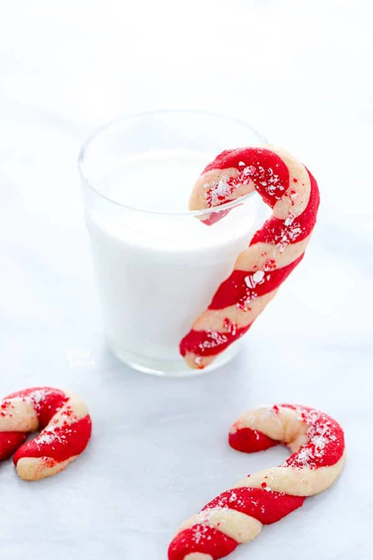 A candy cane shaped cookie hung on a glass of milk.