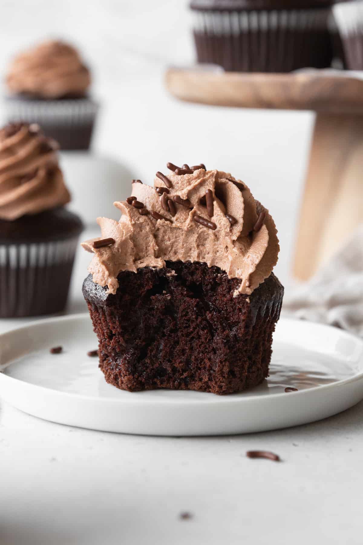 A bitten chocolate cupcake with chocolate frosting on a white plate.