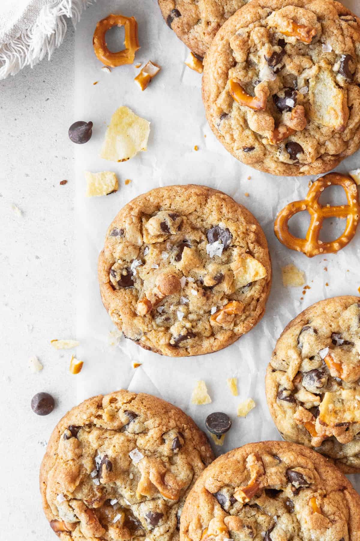 Kitchen sink cookies scattered on a white surface surrounded by chocolate chips, crushed potato chips, and pretzels.