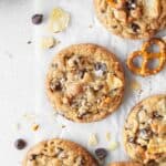 Kitchen sink cookie on a white surface surrounded by more cookies, chocolate chips, crushed potato chips, and pretzels.