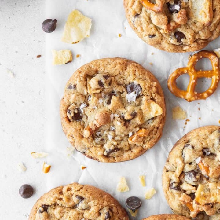 Kitchen sink cookie on a white surface surrounded by more cookies, chocolate chips, crushed potato chips, and pretzels.