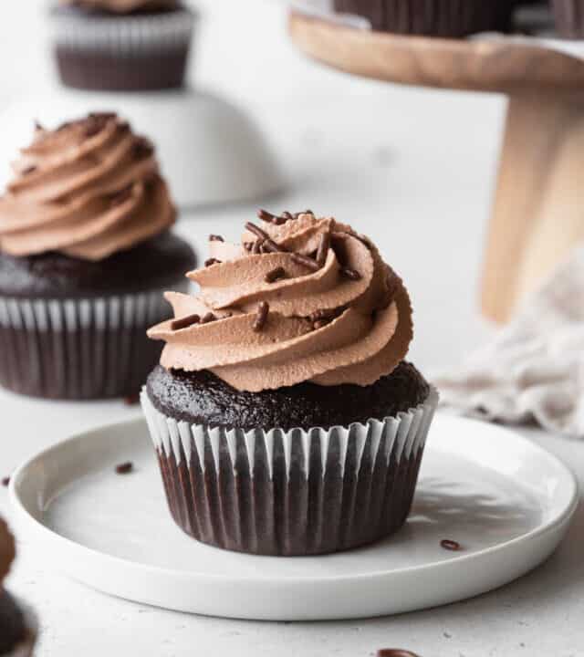 A chocolate cupcake with chocolate frosting on a white plate, with more cupcakes scattered in the background.