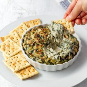 A hand dipping a cracker into a plate of vegan spinach artichoke dip.