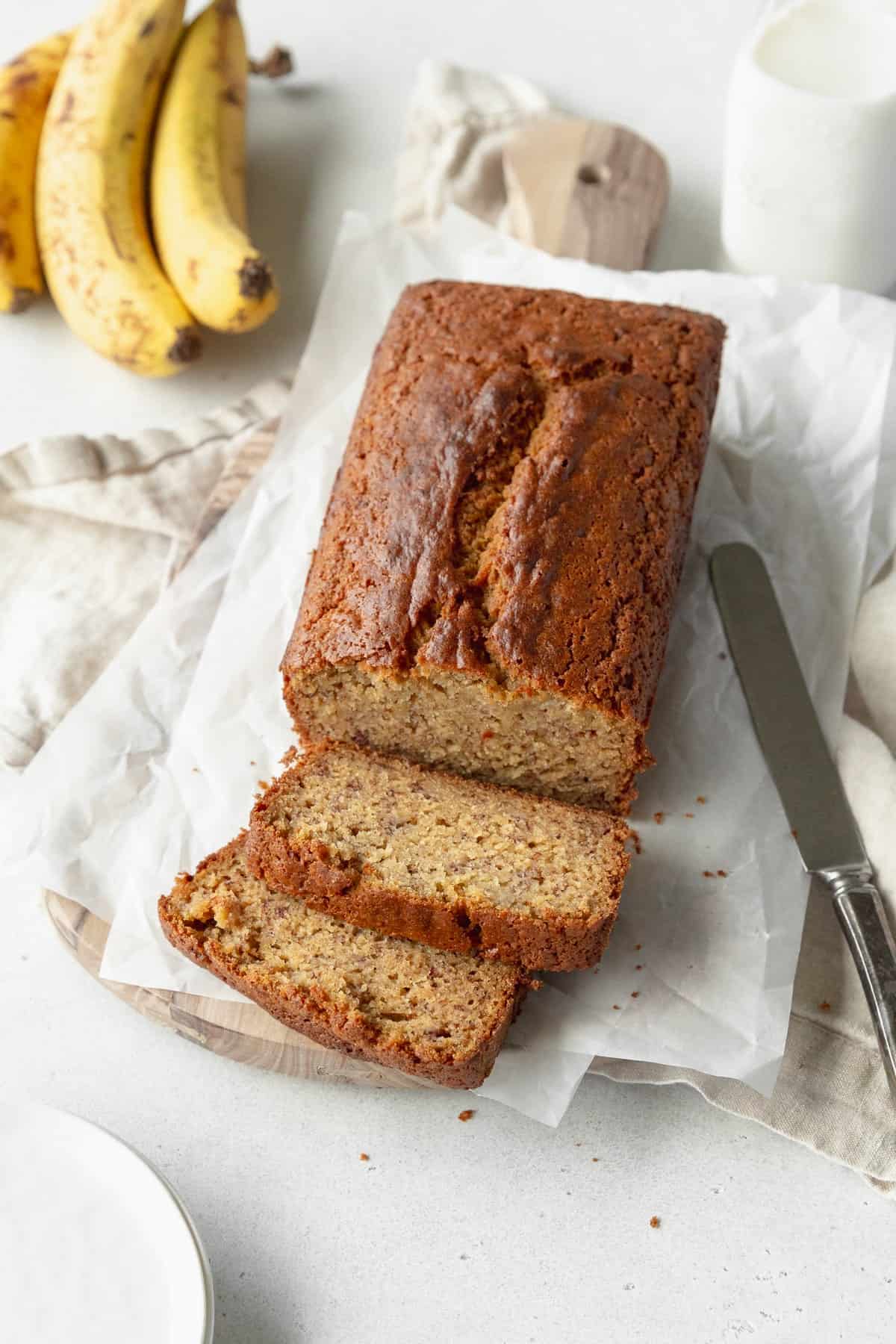 A sliced loaf of gluten free banana bread on sheets of parchment paper with a bunch of banana in the background.