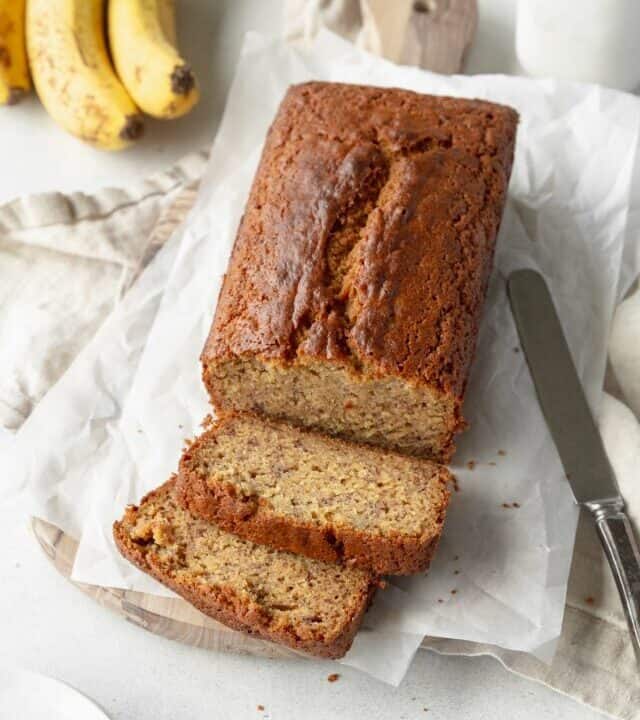 A sliced loaf of gluten free banana bread on sheets of parchment paper with a bunch of banana in the background.