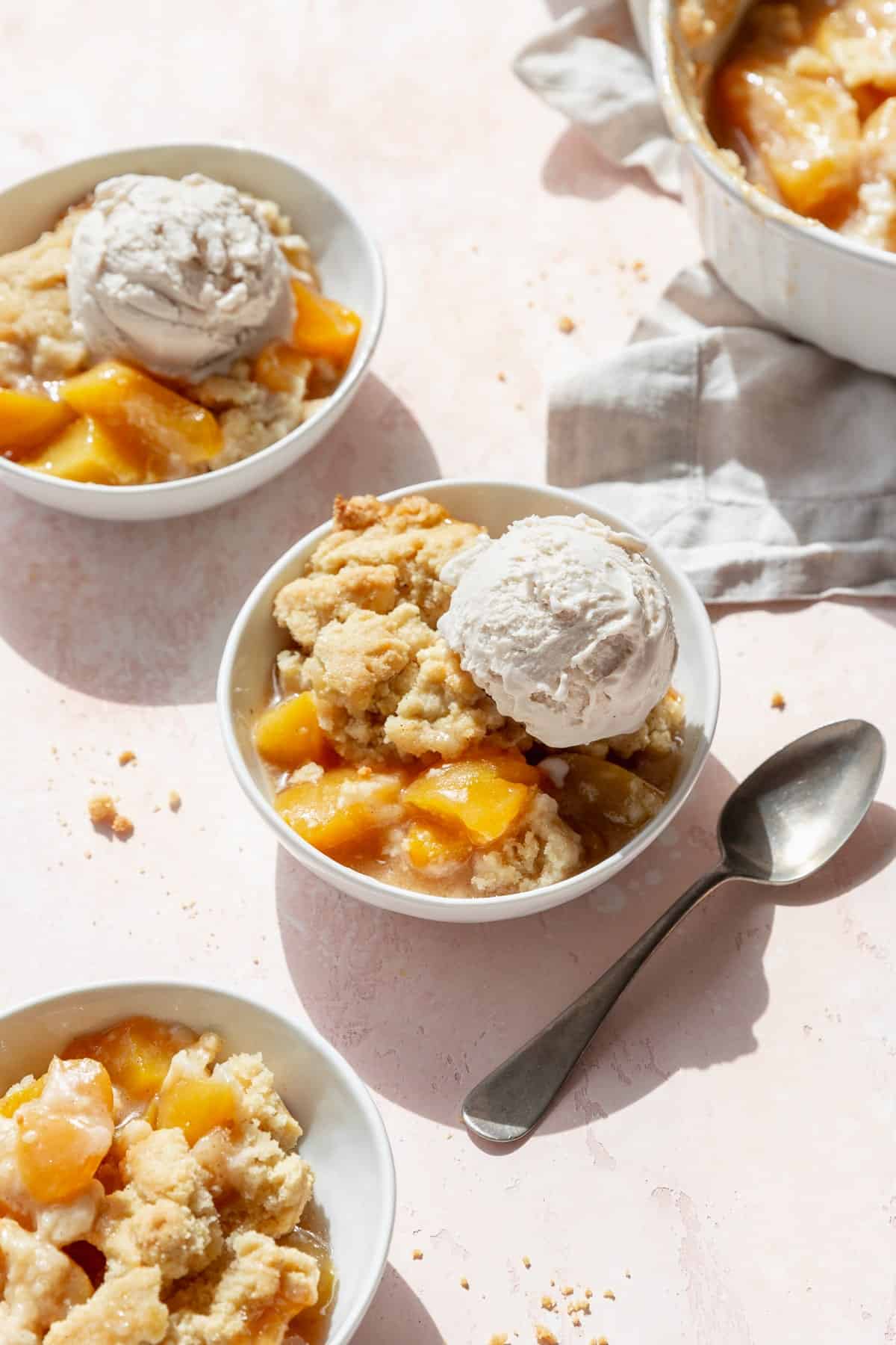 Peach cobbler with a scoop of ice cream in a dessert bowl with a spoon next to it, and more bowls scattered around.