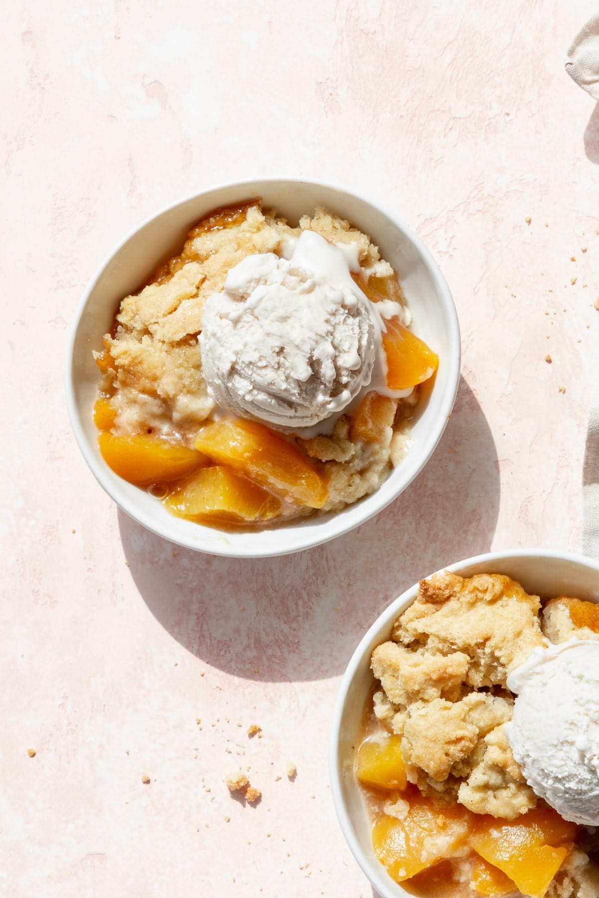 A bowl of peach cobbler with canned peaches and a scoop of vanilla ice cream on a blush colored surface.