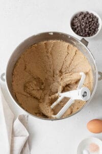 Creamed butter and sugar in the bowl of a stand mixer with an egg on the side and a bowl of chocolate chips in the background.