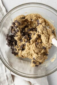 Chocolate chips being folded into almond flour cookie dough in a glass bowl.