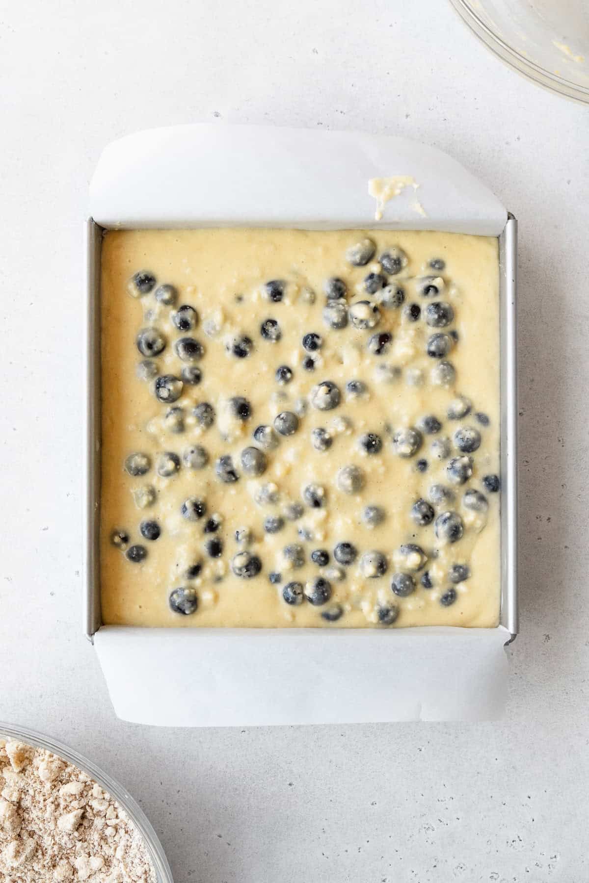 Blueberry cake batter in a square pan on a white surface.