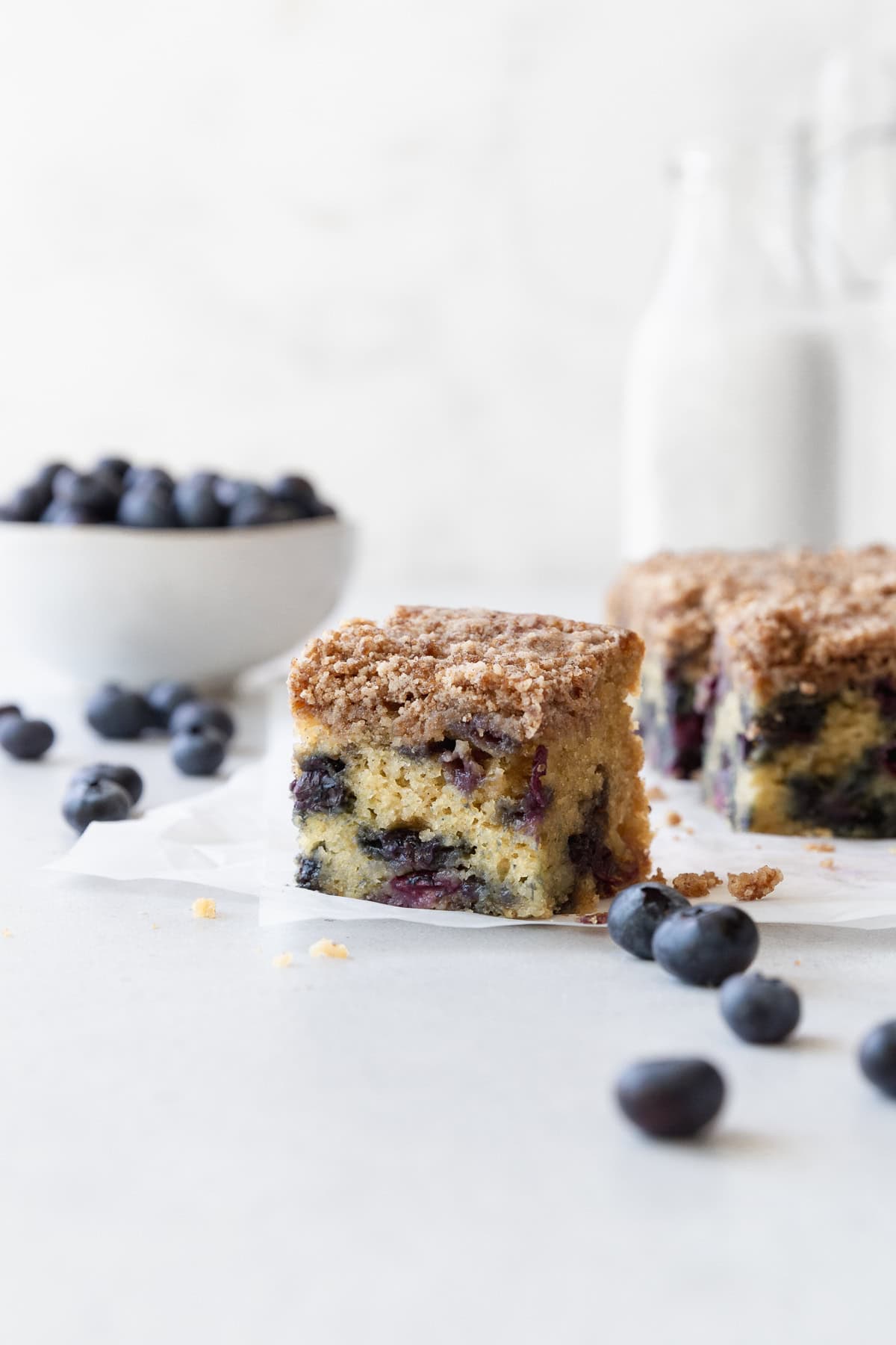 A square of blueberry coffee cake on a white surface with fresh blueberries scattered around.