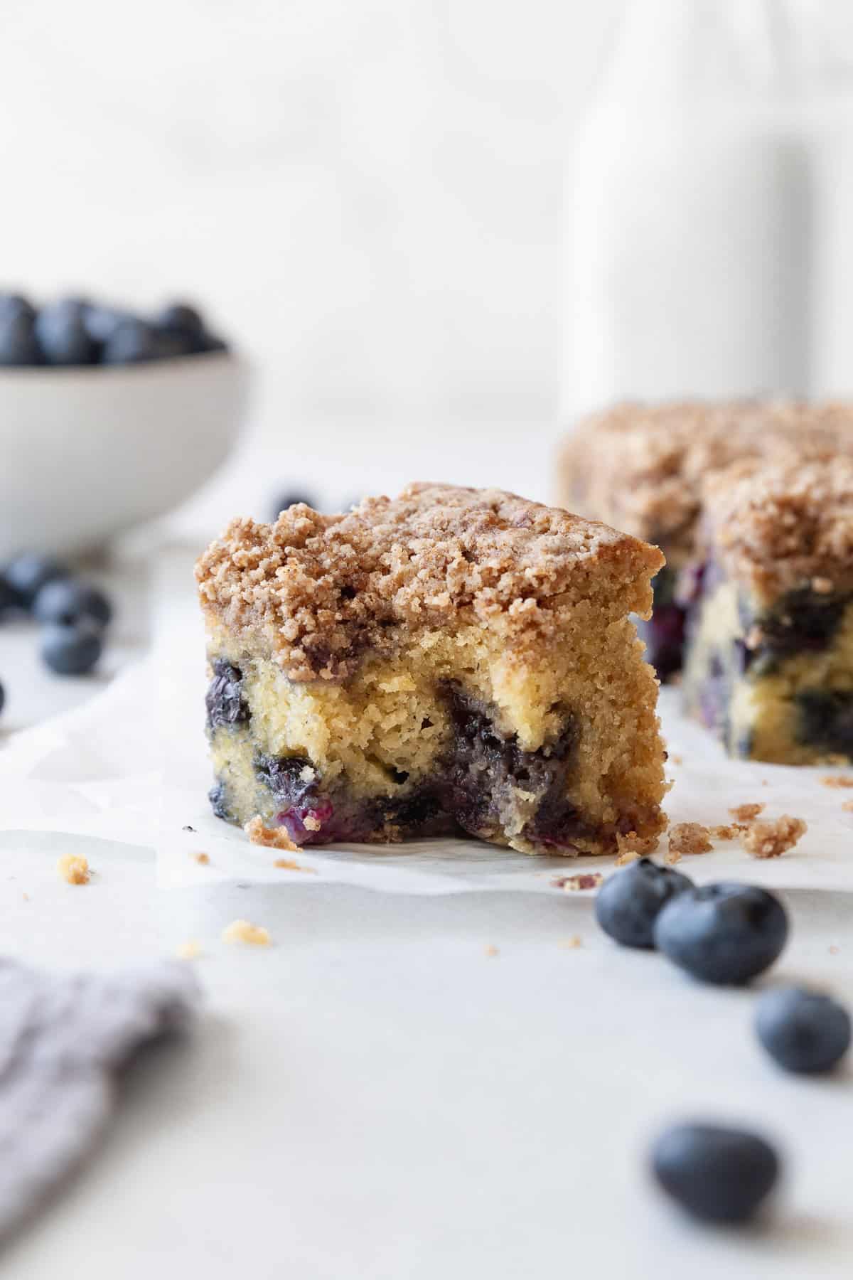 A blueberry coffee cake square with a bite taken out, with a bottle of milk and a bowl of blueberries in the background.