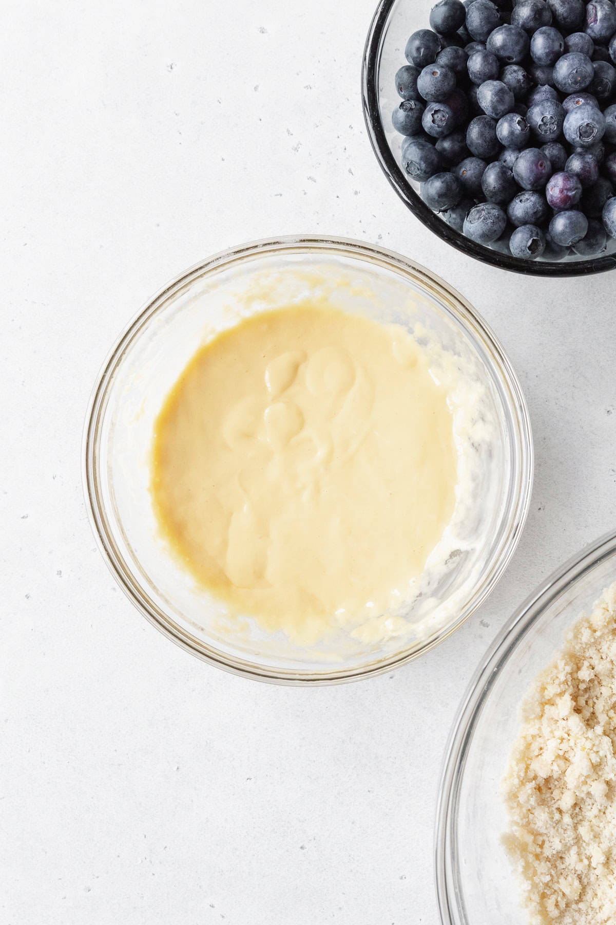 Wet ingredients mixed in a glass bowl with a bowl of blueberries diagonally above it.