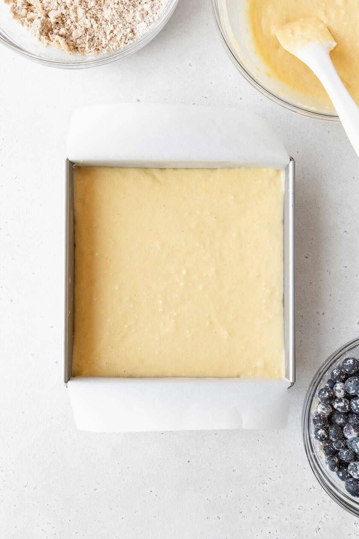 Cake batter in a square pan surrounded by more bowls of ingredients.