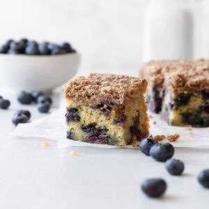 A slice of blueberry coffee cake on a white surface with blueberries scattered.
