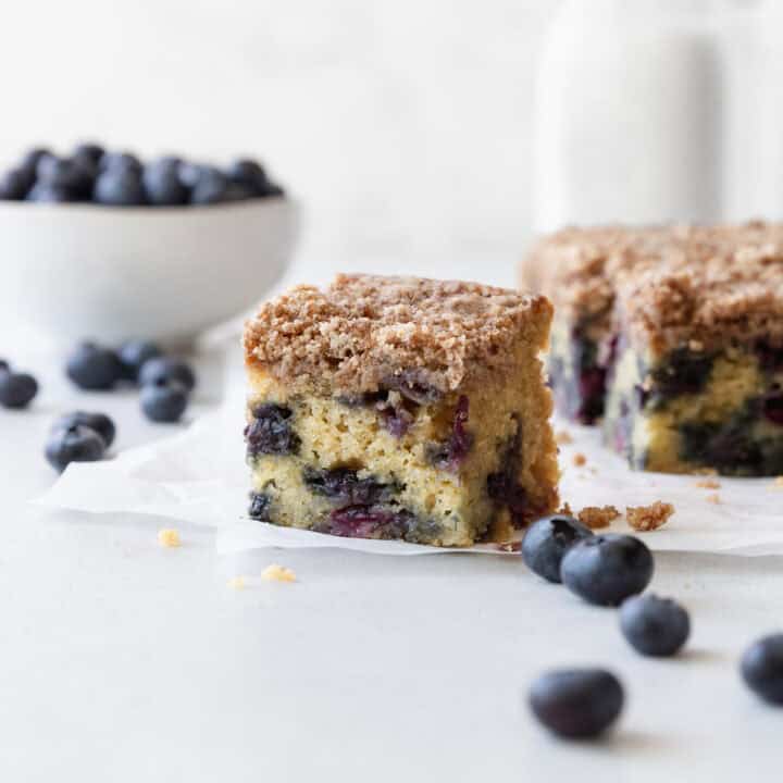 A slice of blueberry coffee cake on a white surface with blueberries scattered.