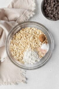 Dry ingredients for oatmeal cookies in a glass bowl next to a natural linen.
